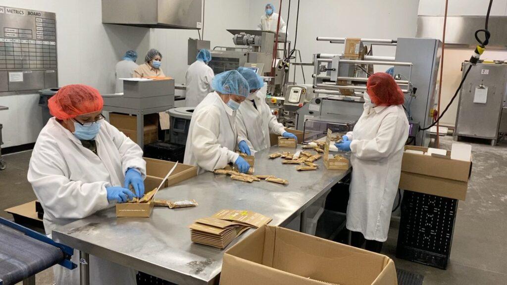 Workers hand-packing product into cartons on the packing line, with the daily KPI metrics board on the wall